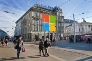 Das NZZ-Gebäude mit Leuten davon, einige Fenster sind mit dem Microsoft-Logo überdeckt. Quellen: NZZ-Gebäude, Falkenstrasse 11 (©NZZ); Microsoft-Logo (©Microsoft). Gesichtsanonymisierung und Collage: Marcel Waldvogel